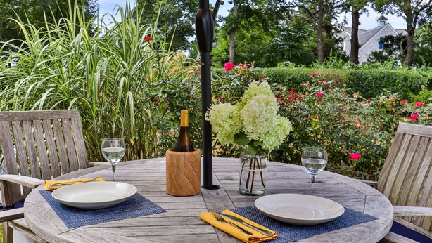 A round wooden table set with plates, cutlery, and wine glasses, surrounded by chairs and a wooden bench. - Unique Oyster Pond 3bd-2ba home in private setting close to downtown. (Chatham)