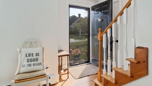 A modern entryway with a wooden staircase, a white chair with a cushion, and a door leading outside. - Unique Oyster Pond 3bd-2ba home in private setting close to downtown. (Chatham)