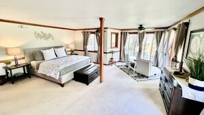 A spacious bedroom with a large bed, a desk, and a chair, a small balcony, and a view of the outdoors.