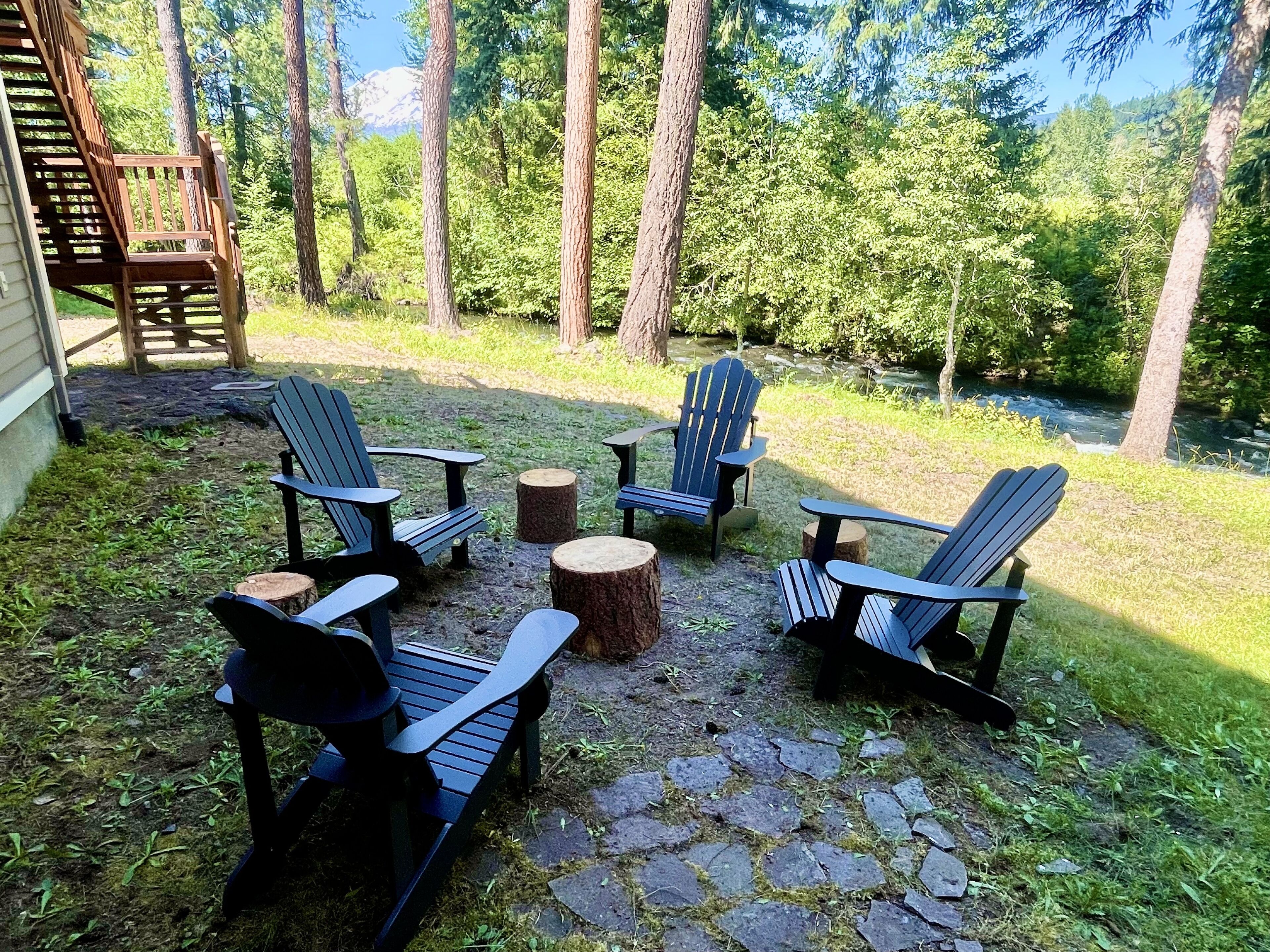 Een terras met Adirondack-stoelen en een stenen tafel, omgeven door een natuurlijke omgeving met bomen en uitzicht op een rivier.