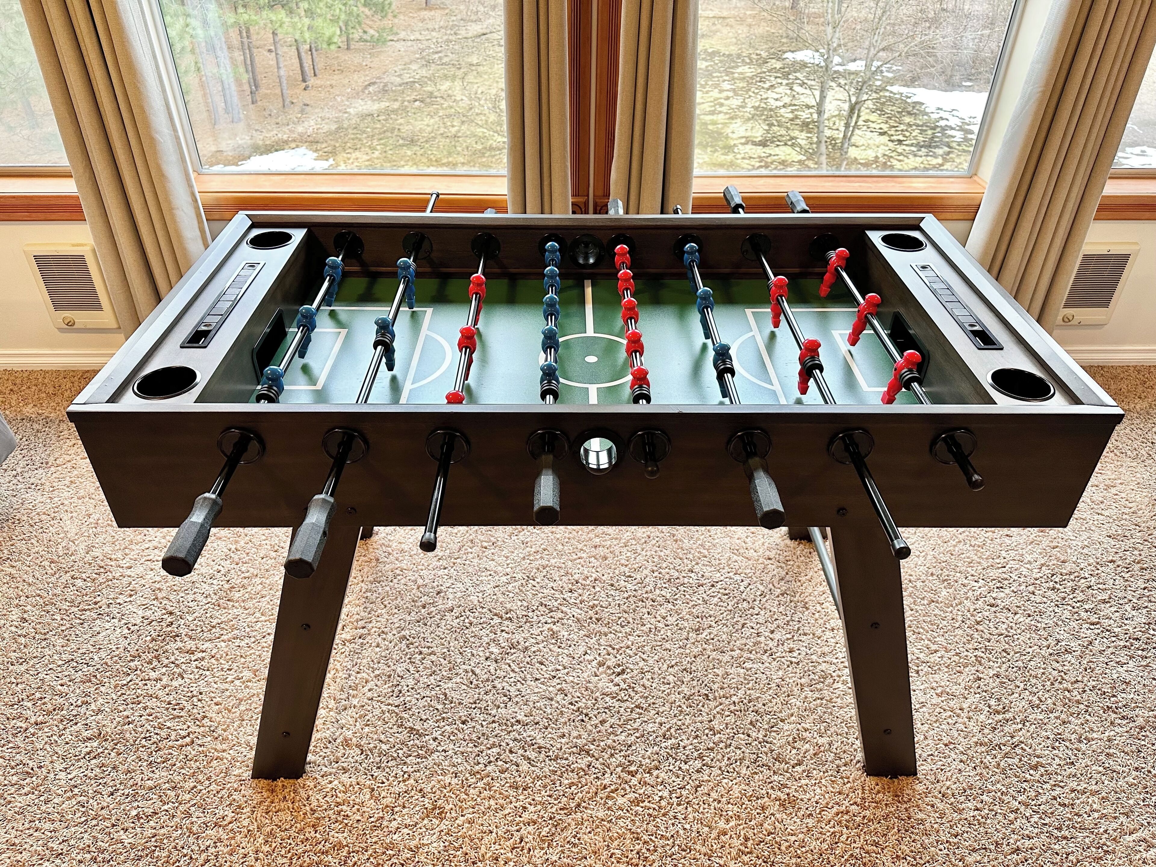 Une table de baby-foot dans une pièce dont le sol est recouvert d’une moquette et qui possède de grandes fenêtres.