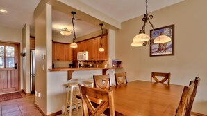 Un vaste espace de salle à manger avec une table et des chaises en bois, une cuisine dotée de meubles en bois et d’un réfrigérateur, ainsi qu’un éclairage suspendu.