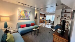 A living room with a sofa, a coffee table, and a TV. There is a kitchen in the background with a dining area.