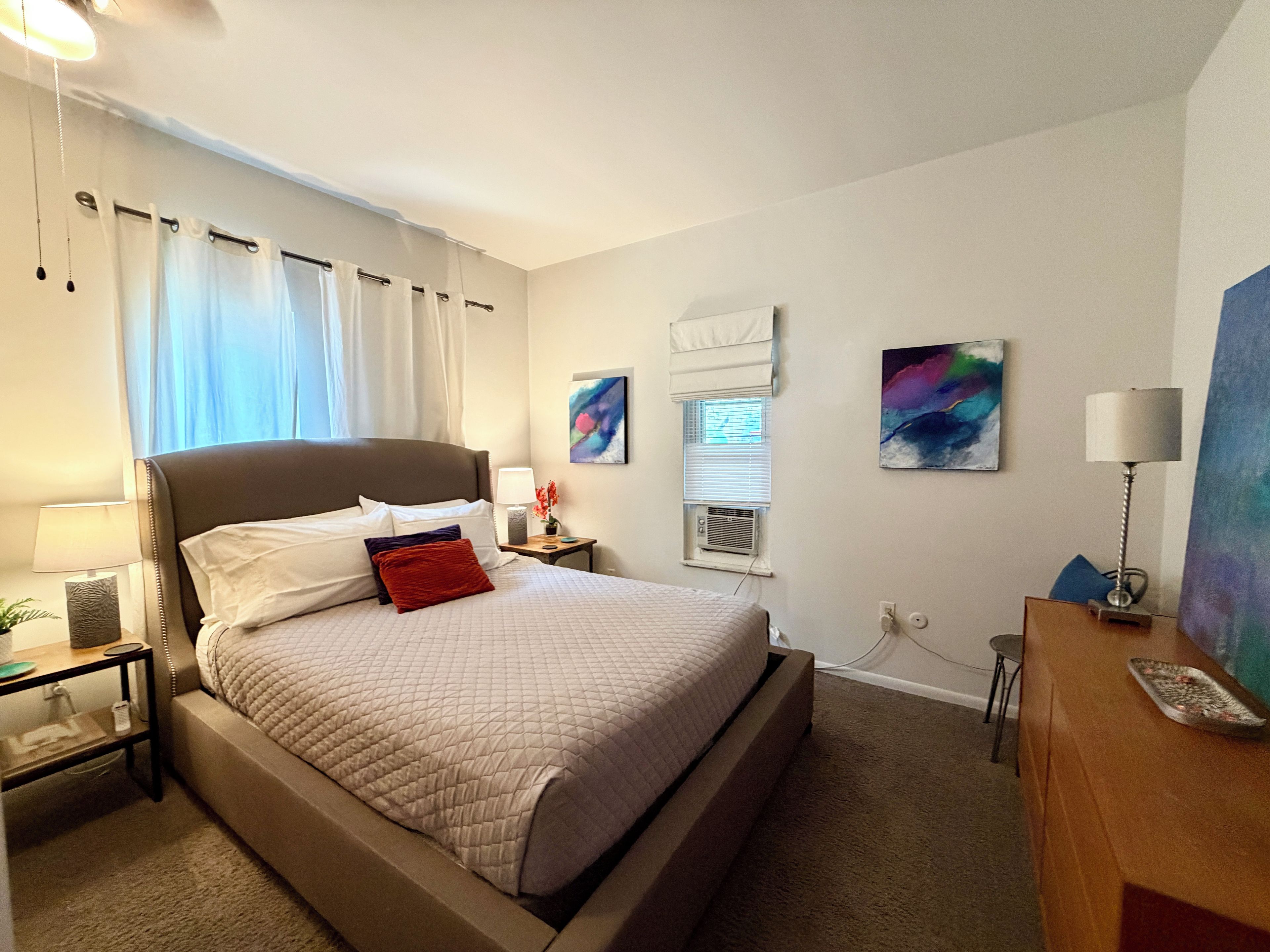 A hotel room with a bed, bedside table, lamp, artwork, and a desk with a lamp.