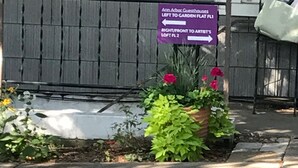 Señal con indicaciones a casas de huéspedes y lofts, una jardinera con flores y una valla de madera.