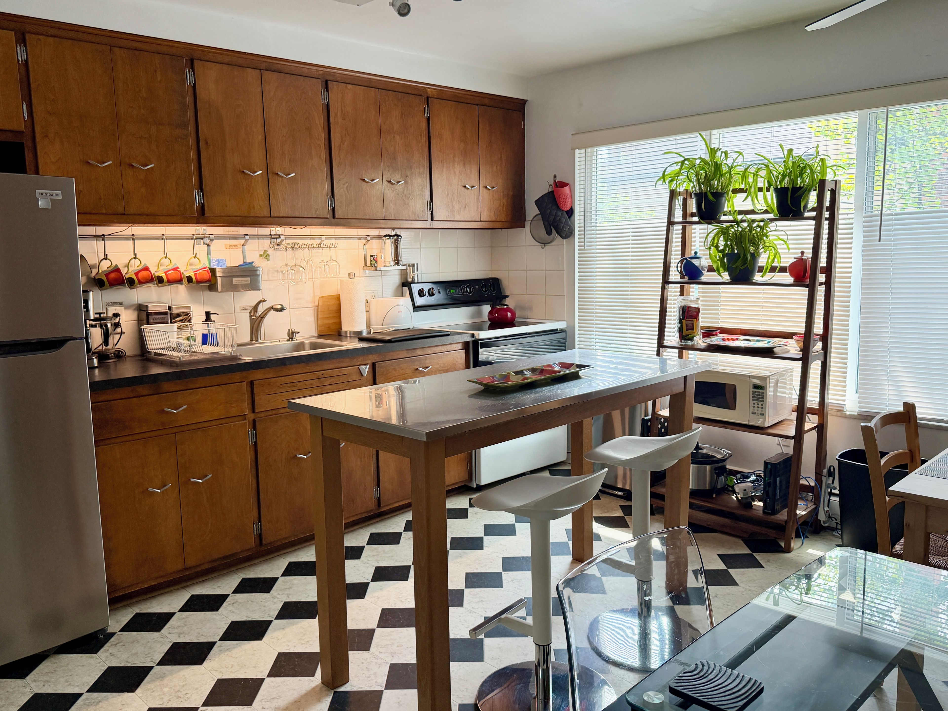 Een keuken met houten kastjes, een roestvrijstalen koelkast, een zwart-wit geruite vloer, een glazen tafel en een raam met planten.