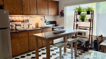 Een keuken met houten kastjes, een roestvrijstalen koelkast, een zwart-wit geruite vloer, een glazen tafel en een raam met planten.