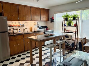 Een keuken met houten kastjes, een roestvrijstalen koelkast, een zwart-wit geruite vloer, een glazen tafel en een raam met planten.