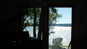 Una habitación con un piano, una silla blanca y vista a un lago con árboles y un bote.