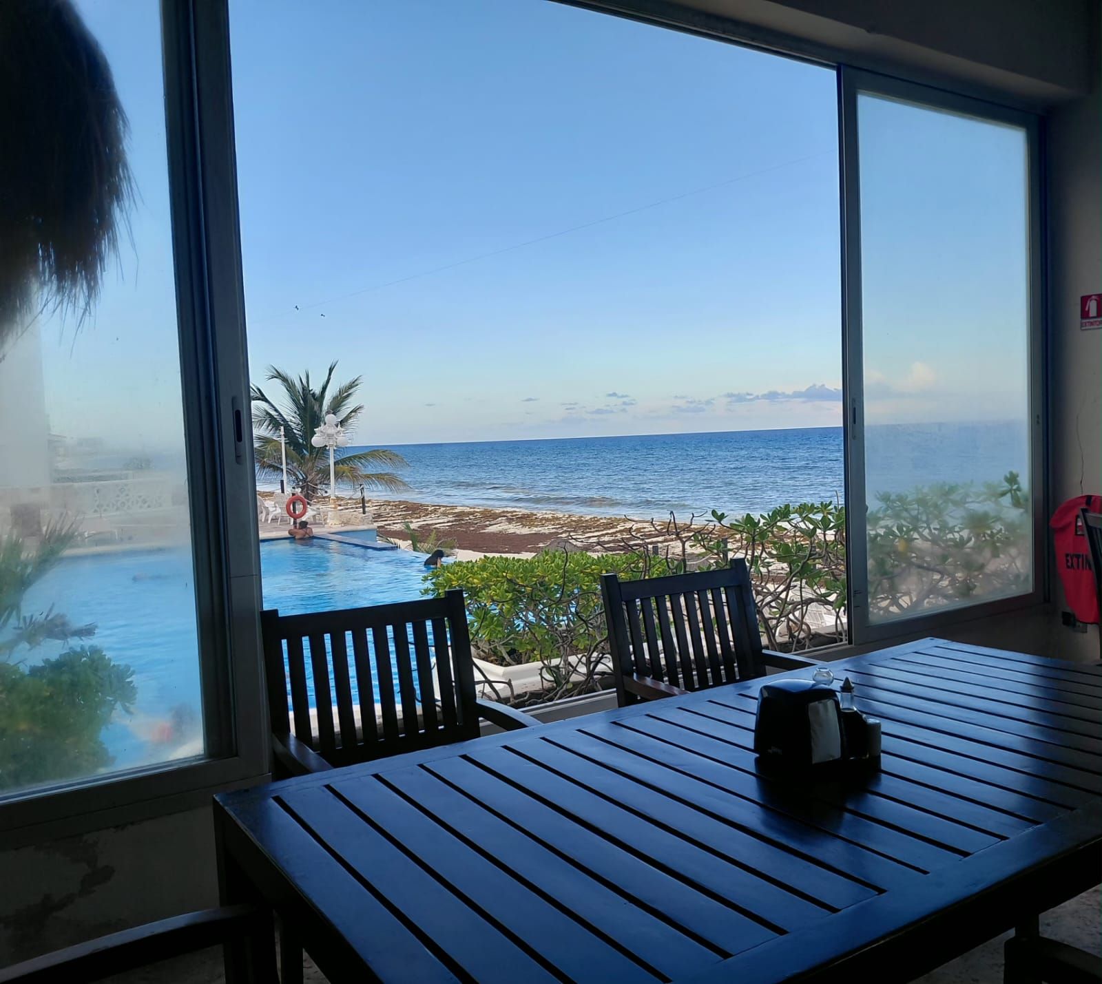 A room with a large window overlooking a pool and beach, a wooden table, and two chairs.