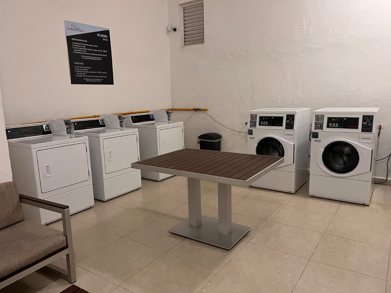 Une buanderie équipée de plusieurs machines à laver, d’une table et d’une chaise. Des instructions sont affichées sur le mur.