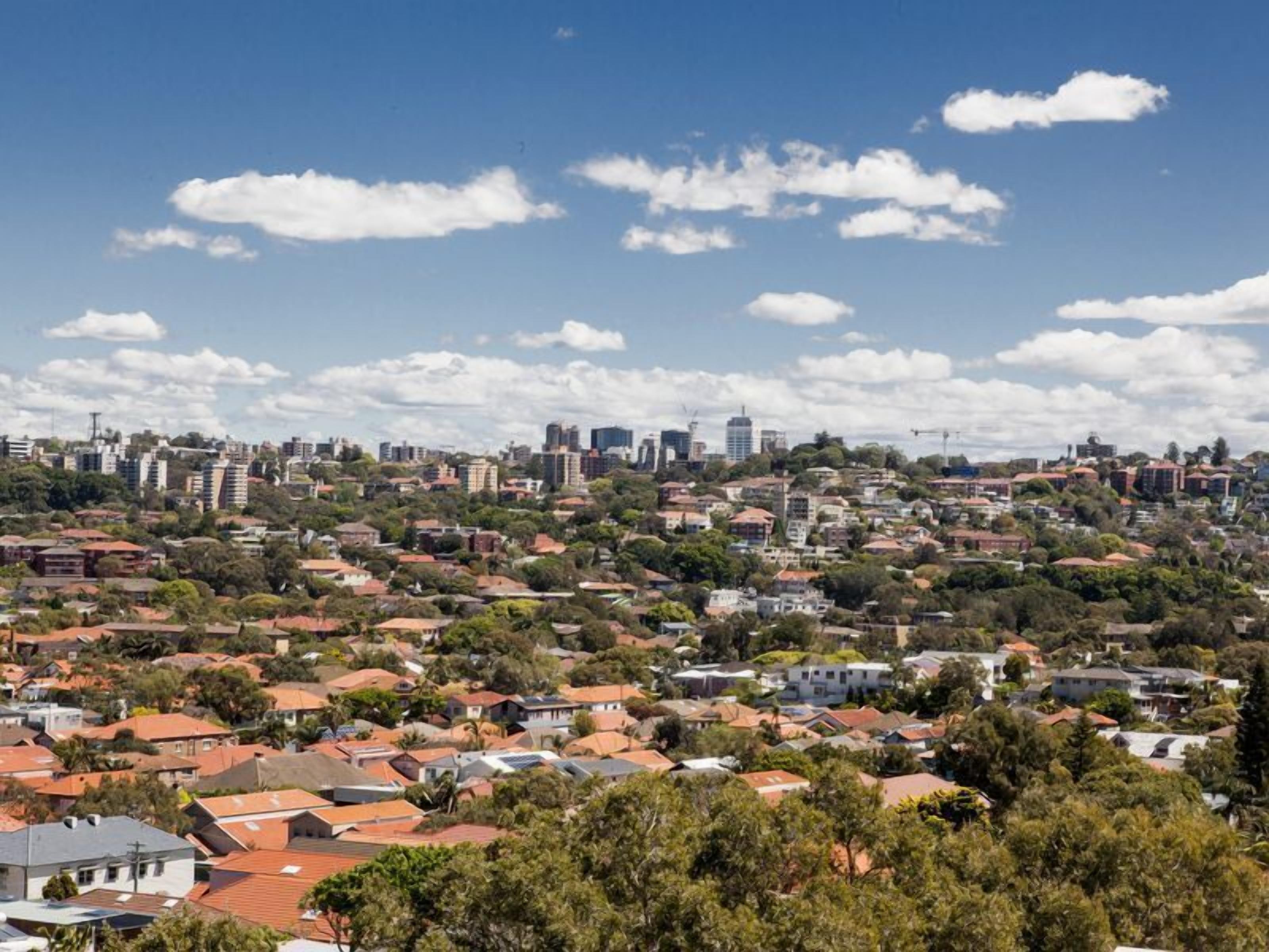 Zona residencial amb cases i edificis diversos, envoltada de vegetació i sota un cel blau.