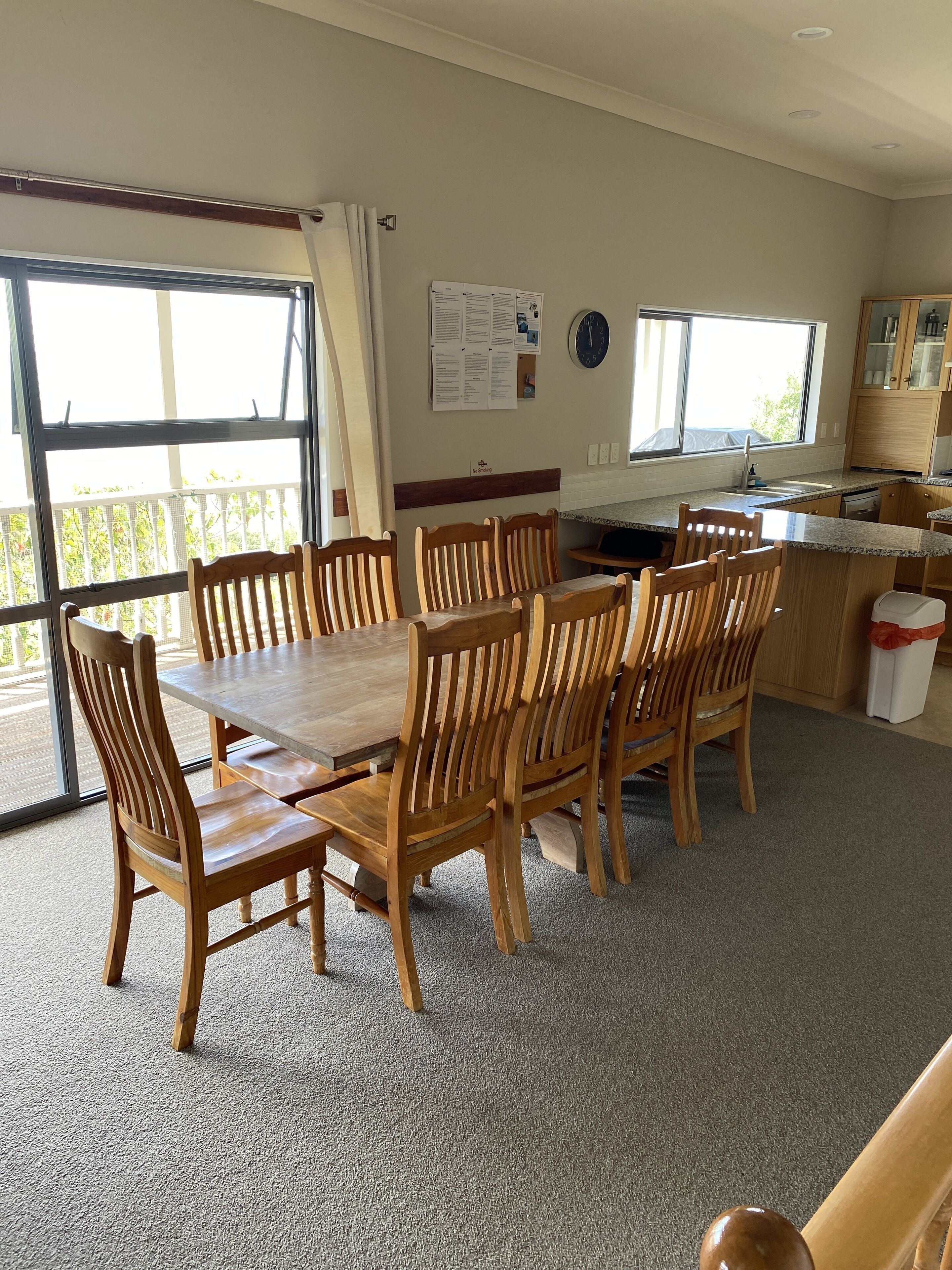 A dining area with a wooden table and chairs, a kitchen area with cabinets and a sink, and a view through large windows.