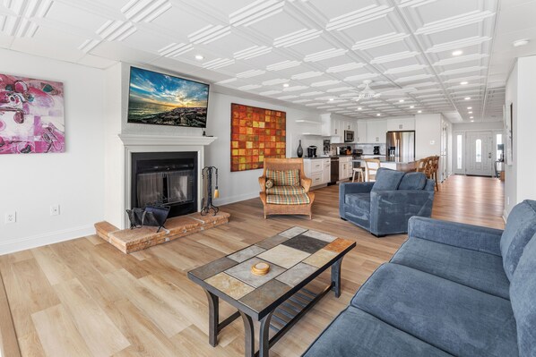 A spacious living room with a fireplace, a large painting, and a modern kitchen in the background.
