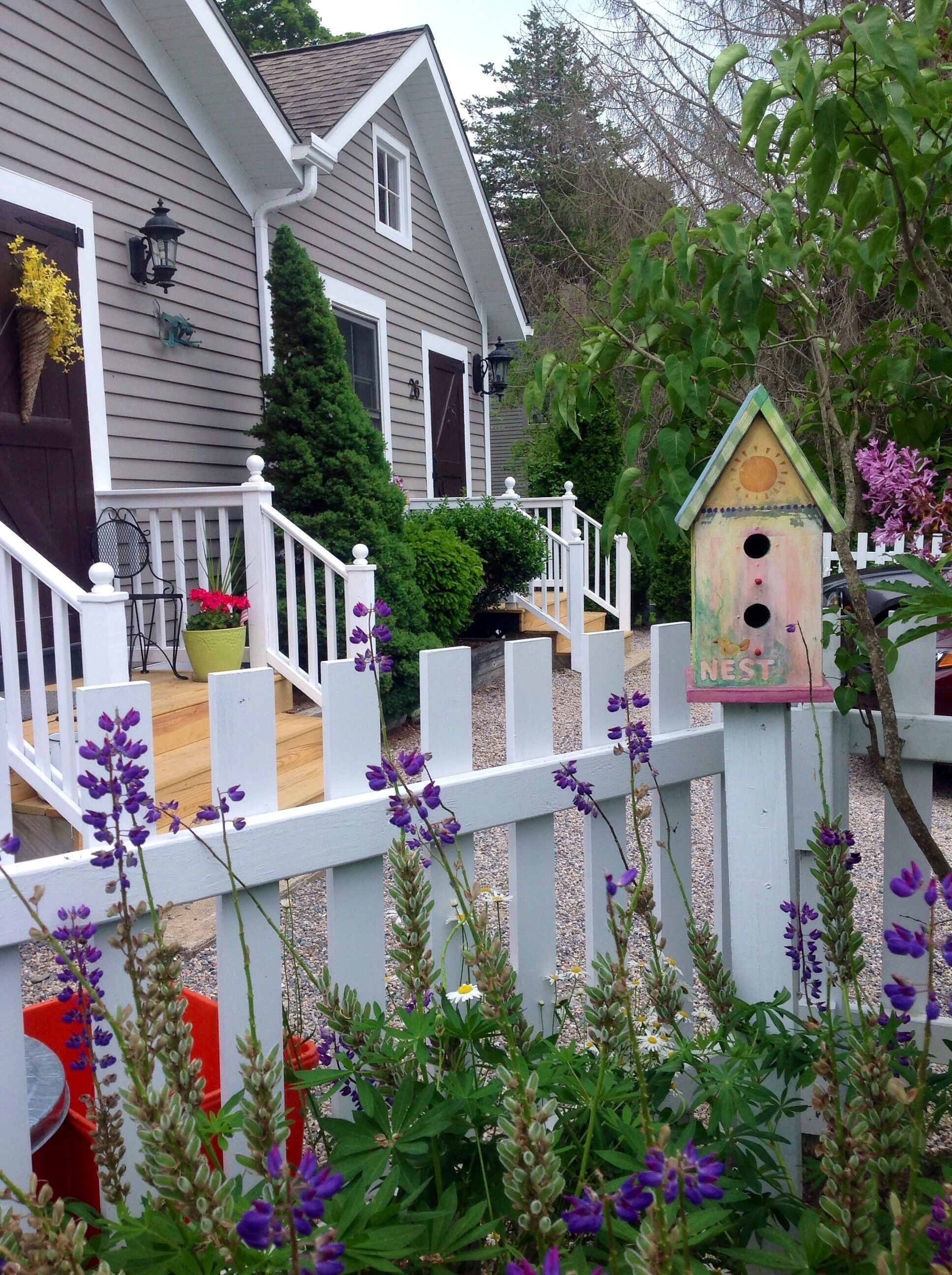 Une maison avec une clôture blanche en bois, un nichoir et des fleurs violettes.