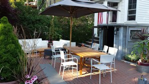 A patio with a wooden table and white wicker chairs, surrounded by potted plants and a large umbrella. - Charming Tree Top Victorian - Two Bedroom Retreat (Boston)