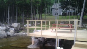 A wooden dock with a picnic table and benches, overlooking a body of water with rocks and trees in the background. - Elegant, spacious 5 bedroom home on Molasses Pond (Eastbrook)