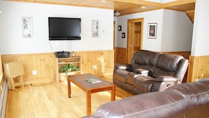 A living room with wooden paneling, a leather sofa, a coffee table, a flat-screen TV, and a wooden cabinet. - Elegant, spacious 5 bedroom home on Molasses Pond (Eastbrook)