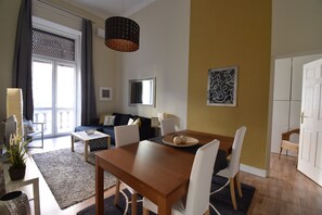 Ein modernes Wohnzimmer mit Essbereich, ausgestattet mit einem Holztisch, weißen Stühlen, einer Couch und einem großen Fenster mit Vorhängen.