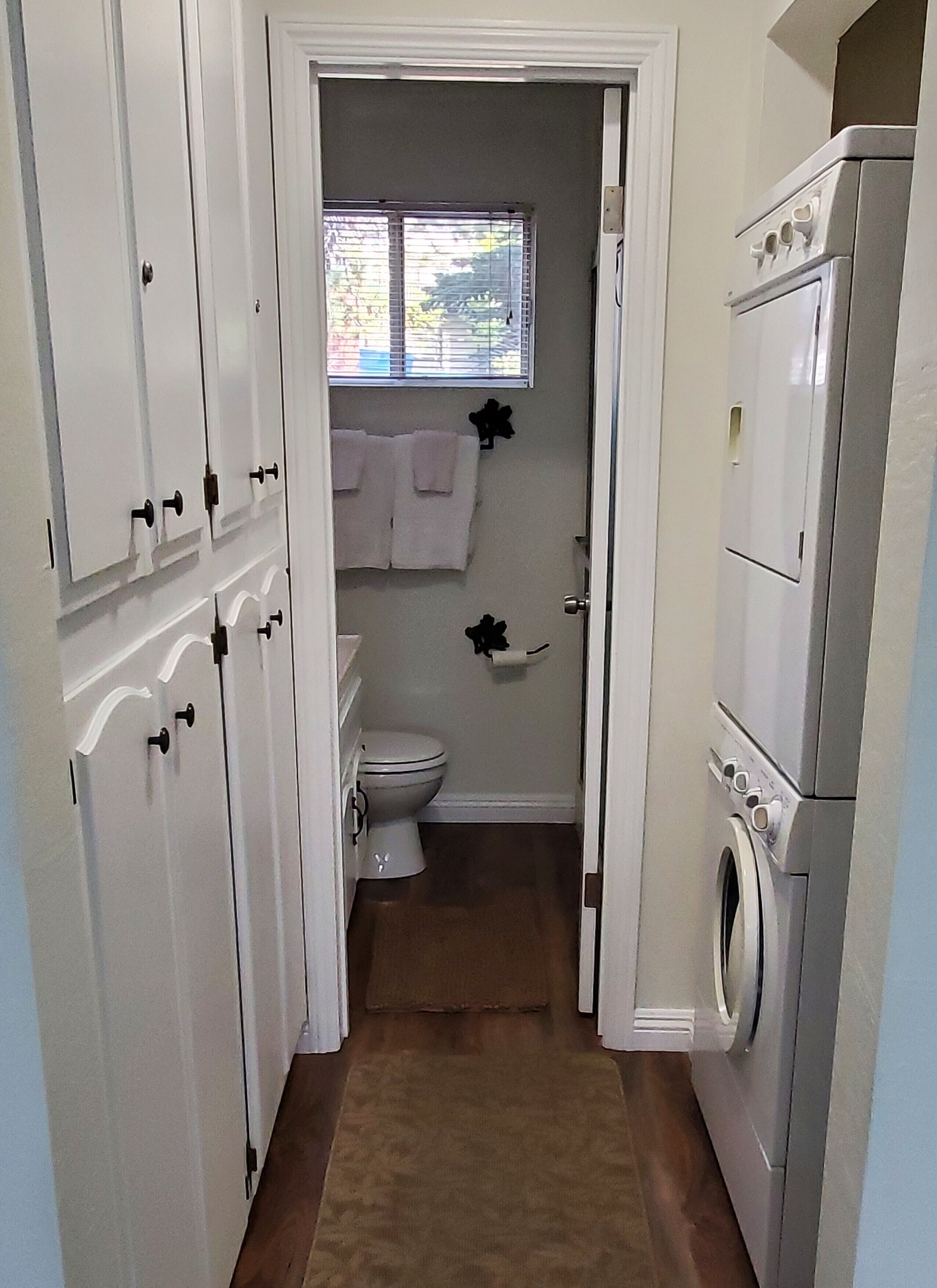 Een smalle gang die leidt naar een badkamer met een toilet, een raam met zonwering en een wasmachine.