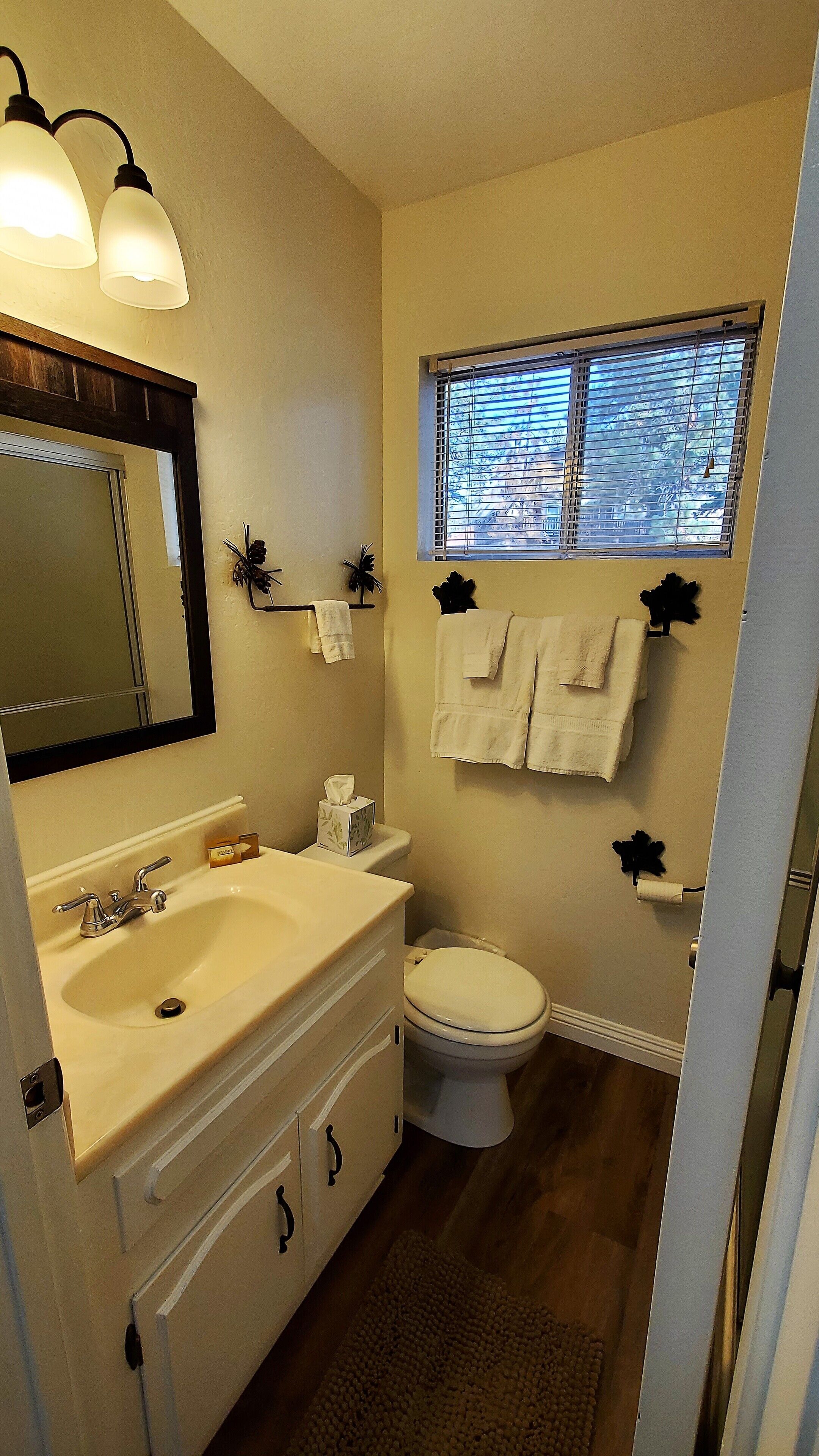 A bathroom with a sink, mirror, and toilet. There are towels hanging on the wall and a window above the toilet.