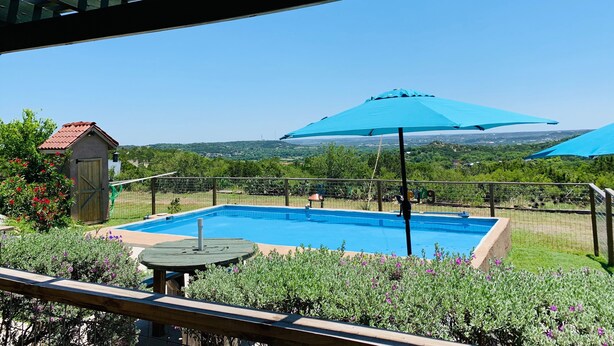 A swimming pool with a wooden deck, surrounded by a fence and covered by a blue umbrella. - Beautiful view,18 acres,pool, game room,Axis deer, 3 mile’s to the  river trail! (Kerrville)