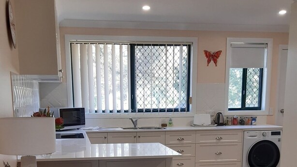 A modern kitchen with a central island, a built-in washing machine, and a dining area with a wooden table and chairs. - Butterfly Cottage - Pet Friendly Tumut (Tumut)