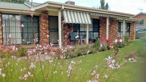A two-story house with a brick facade, a covered porch with chairs, and a lawn with flowering plants. - Butterfly Cottage - Pet Friendly Tumut (Tumut)