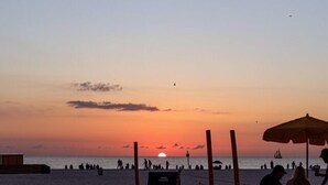 Cena de praia com um pôr do sol, pessoas caminhando e guarda-sóis de praia.
