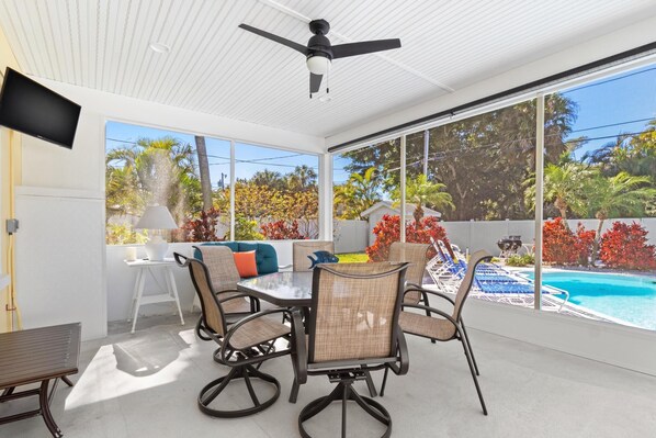 Une terrasse ensoleillée avec une table à manger, des chaises et un ventilateur de plafond. On aperçoit une piscine à travers de grandes fenêtres.