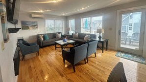 A spacious living room with hardwood flooring, a gray sofa, a coffee table, and two armchairs. There are two windows with blinds, a television mounted on the wall, and a door leading outside.
