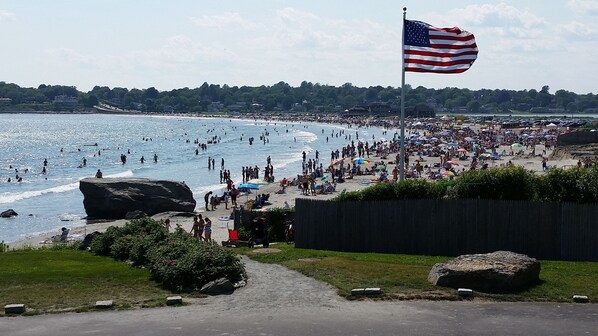 A crowded beach with a large American flag, rocky outcrops, and a path leading to the beach. - Surfside! Fantastic Beach House, Near Restaurants, Cliff Walk, Close to Downtown (Middletown)