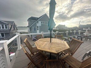 A wooden deck with a round table and chairs, overlooking a beach and houses. - Surfside! Fantastic Beach House, Near Restaurants, Cliff Walk, Close to Downtown (Middletown)