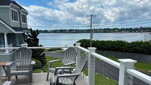 A deck with four Adirondack chairs and a small table, overlooking a lake with a distant shoreline.