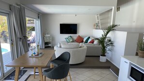 A modern living room with a sofa, a dining table with chairs, a TV mounted on the wall, and a kitchen area with a refrigerator and microwave.