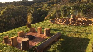 An outdoor seating area with wooden stools and a fire pit, surrounded by a grassy landscape and trees.