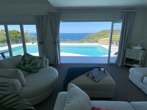 Ampla sala de estar com uma grande janela com vista para uma piscina e para o oceano.