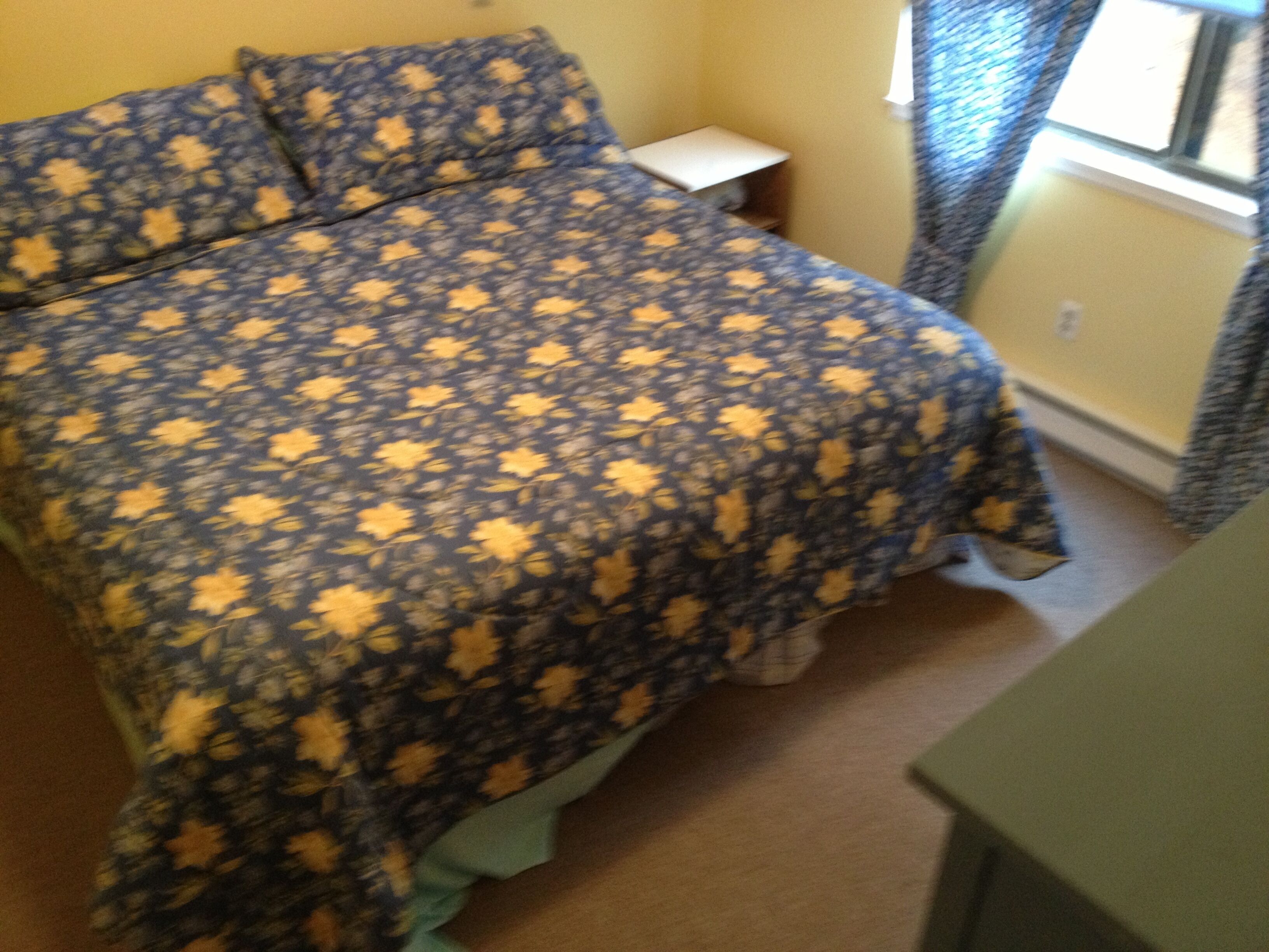 A bed with a floral-patterned bedspread, a small bedside table, and a window with curtains.