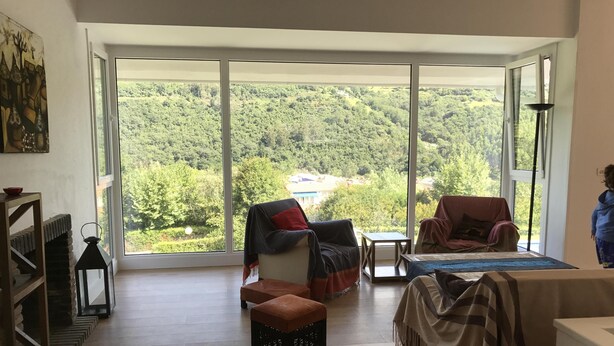Il s’agit d’une pièce spacieuse dotée d’une grande porte-fenêtre coulissante qui donne sur un paysage verdoyant luxuriant. La pièce contient un canapé, un fauteuil et une petite table.