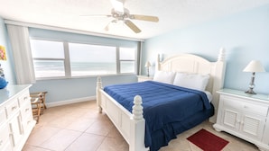 Quarto com uma janela grande, cama com colcha azul, cômoda branca e ventilador de teto.