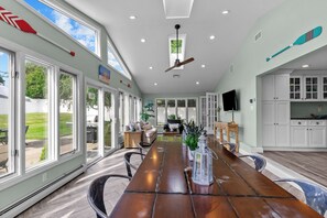 Une salle à manger spacieuse avec une longue table en bois, de grandes fenêtres et un ventilateur de plafond.
