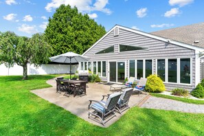 Une maison moderne dotée d’un patio comprenant un barbecue, du mobilier d’extérieur et une grande véranda avec de larges fenêtres.