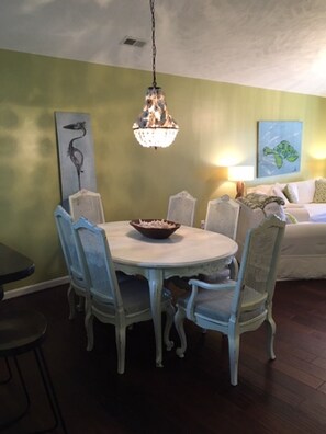 A dining area with a round table, six chairs, a chandelier, a painting of a bird, and a sofa with pillows.