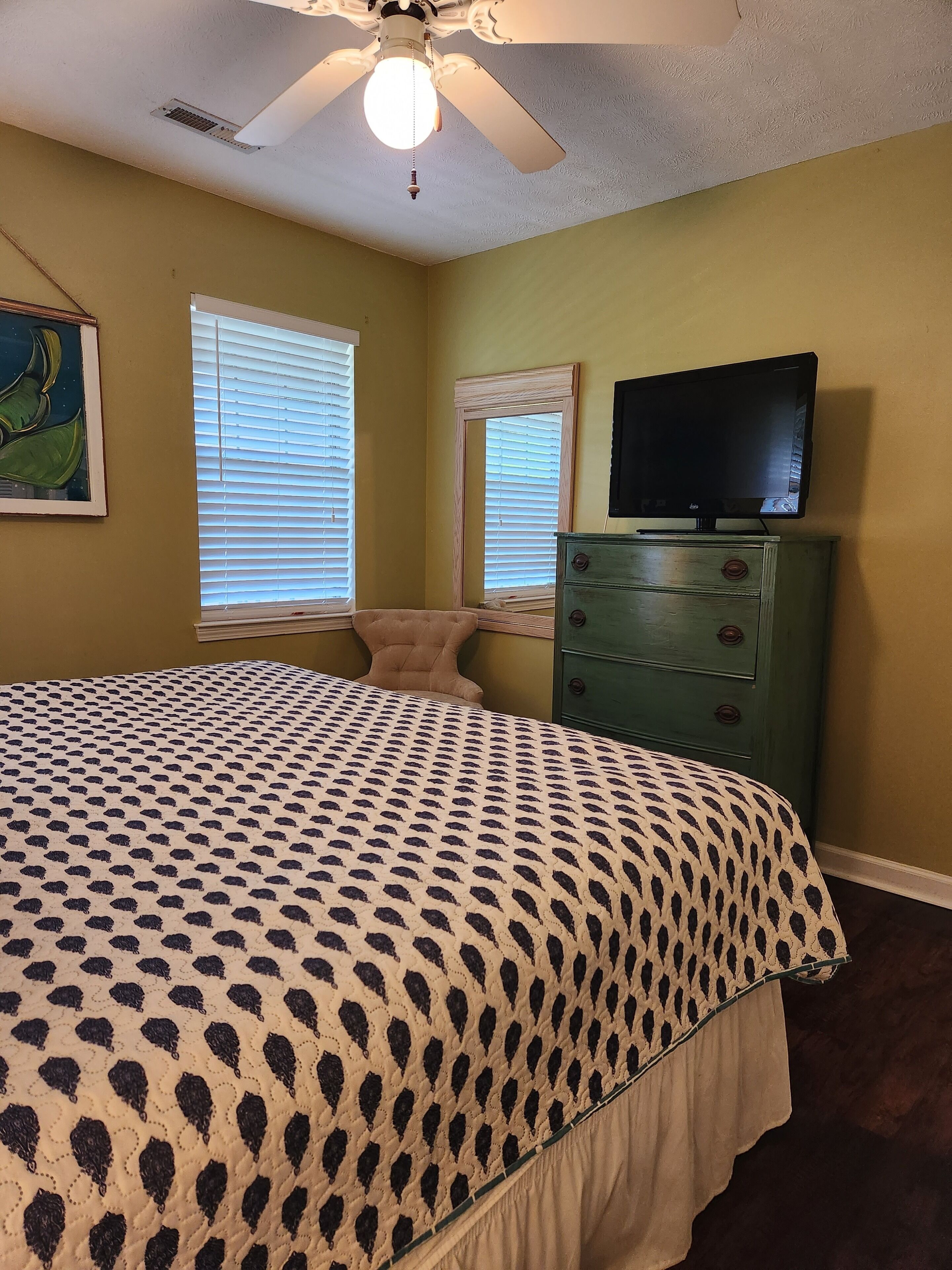 A bedroom with a bed, a dresser, a television, and two windows with blinds.