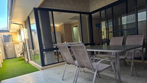En udendørs terrasse med et bord og stole, en glasdør der fører ind til et værelse, og en lille græsplæne.