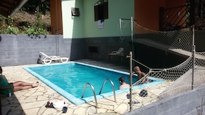 Piscina com piso de pedra, rede de segurança com corda e uma pequena janela no prédio.