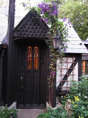 A black wooden door with intricate designs, a small garden with various plants, and a stone statue. - Romantic Herringbone Cottage - In Our Little English Village (New Hope)