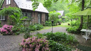 Uma casa rústica com fachada de tijolos e telhado inclinado, rodeada por vegetação exuberante e plantas floridas.