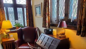 Quarto com sofá de couro, uma cadeira de madeira, uma pequena mesa com luminária, um rádio e uma vela. Há janelas com cortinas e um quadro na parede.