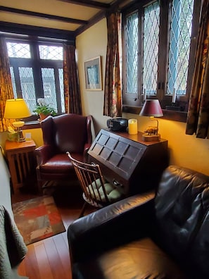 Quarto com sofá de couro, uma cadeira de madeira, uma pequena mesa com luminária, um rádio e uma vela. Há janelas com cortinas e um quadro na parede.